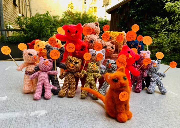 En hel flok hæklede katte i røde, orange og lyserøde farver står i en gård med små runde skilte på pinde.