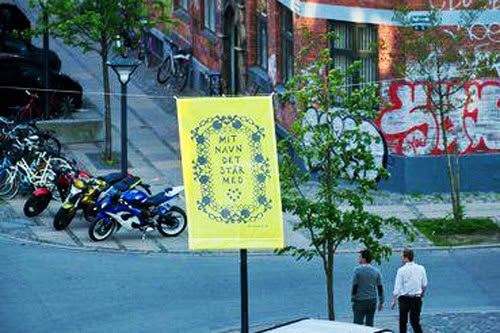Et stort gult banner med et sort gækkebrevsklip hænger på en lygtepæl ved et gadehjørne med røde murstenshuse.