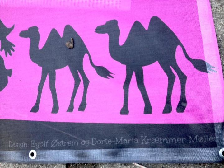 Hjørnet af et pink banner, der ligger på jorden: benene af to sorte kameler i silhuet og teksten Design: Eyolf Østrem og Dorte-Maria Kræmmer Møller.
