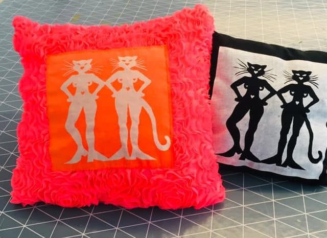 To puder: en lakrød plyspude med orange felt og to sølvfarvede kattedamer, og en hvid pude med to sorte kattedamer.