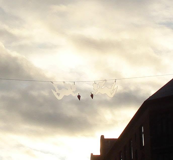 To hvide klippede engle med røde hjerter hænger på en line højt over gaden mod en skyet himmel.