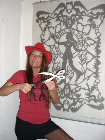Dorte-Maria i rød cowboyhat og rød T-shirt klipper med en stor saks foran et banner med sit eget papirklip, Damen med Saksen.