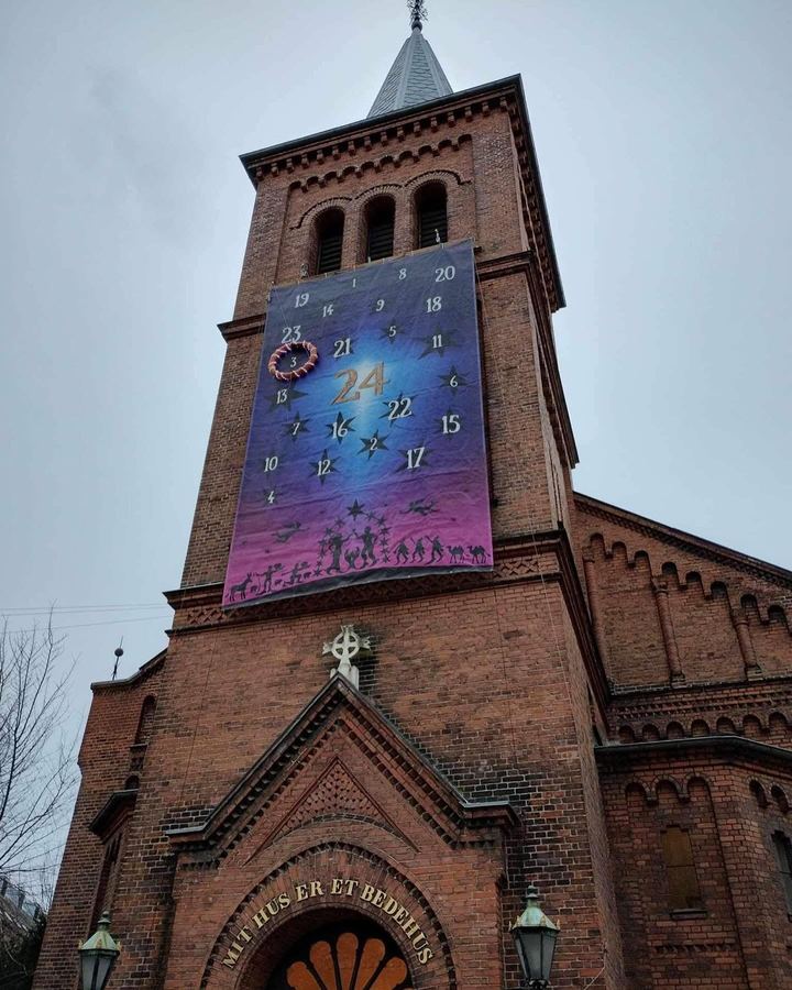 Et stort blåt og pink banner på et rødt kirketårn: tallene 1 til 24 mellem stjerner, en ring om tallet 3, og nederst sorte papirklip af den hellige familie under en stjernebue, hyrder, konger, kameler, engle og et æsel.